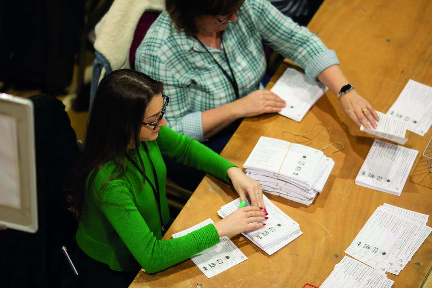 Two women sitting at a large wooden table counting election votes
