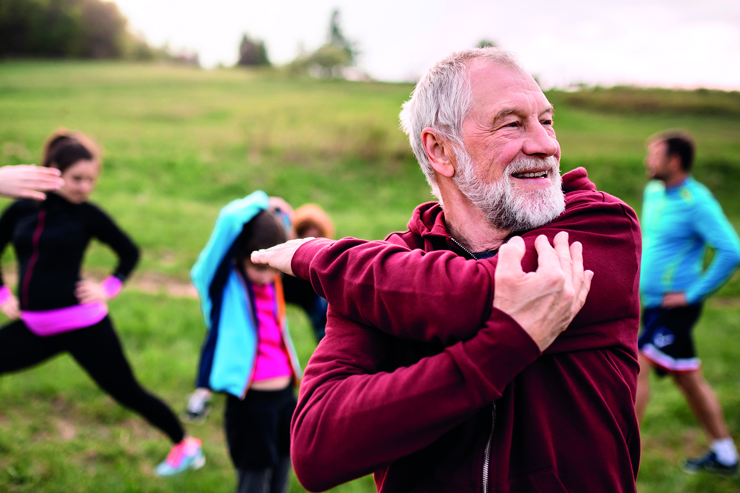 photo of older man stretching his shoulder with others warming up in the background outdoors