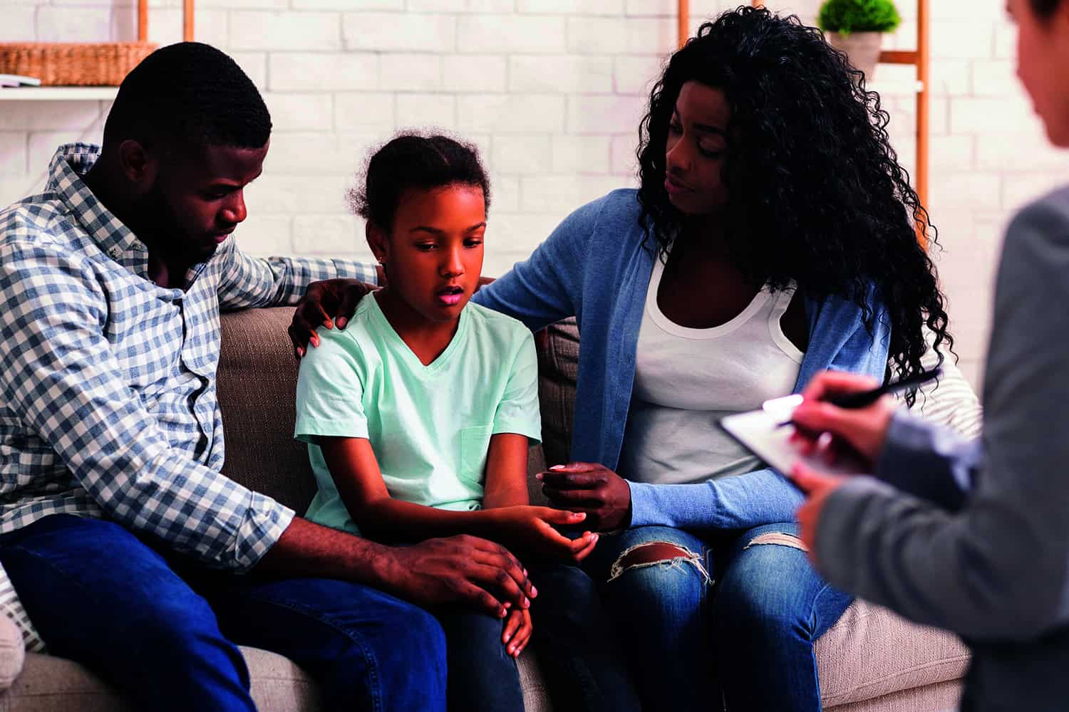 A sad looking child sitting on a sofa with her parents comforting her. A person is listening and writing in a notepad.