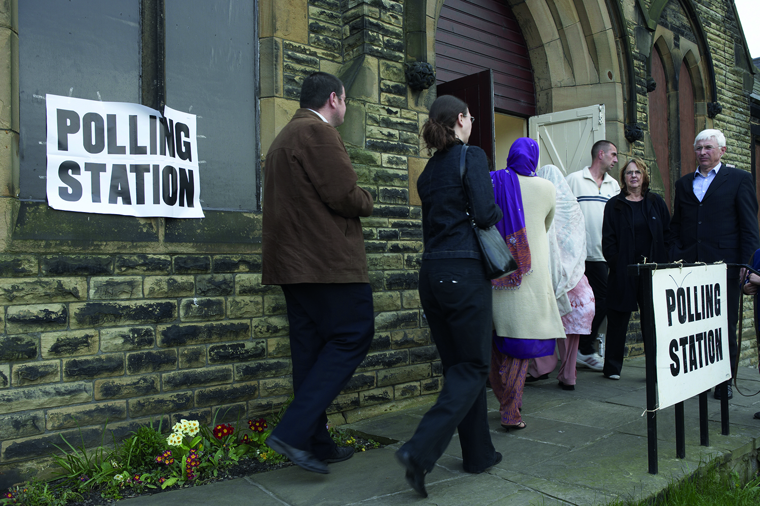A crowd of people entering a polling station, situated within a local church. There are signs saying polling station on the outside