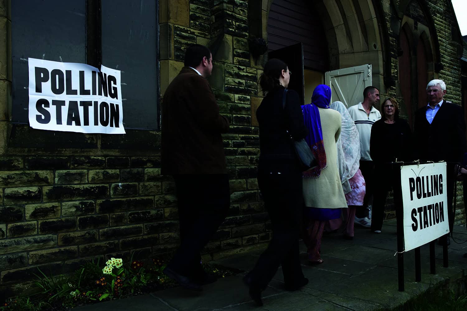 A crowd of people entering a polling station, situated within a local church. There are signs saying polling station on the outside