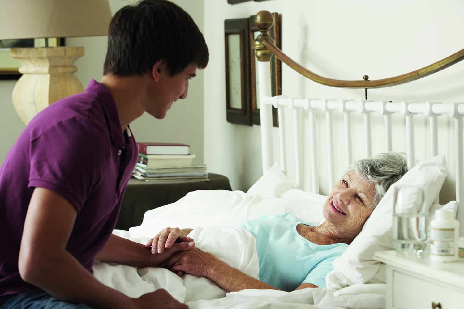 An elderly woman lying in bed receiving help from a young male carer