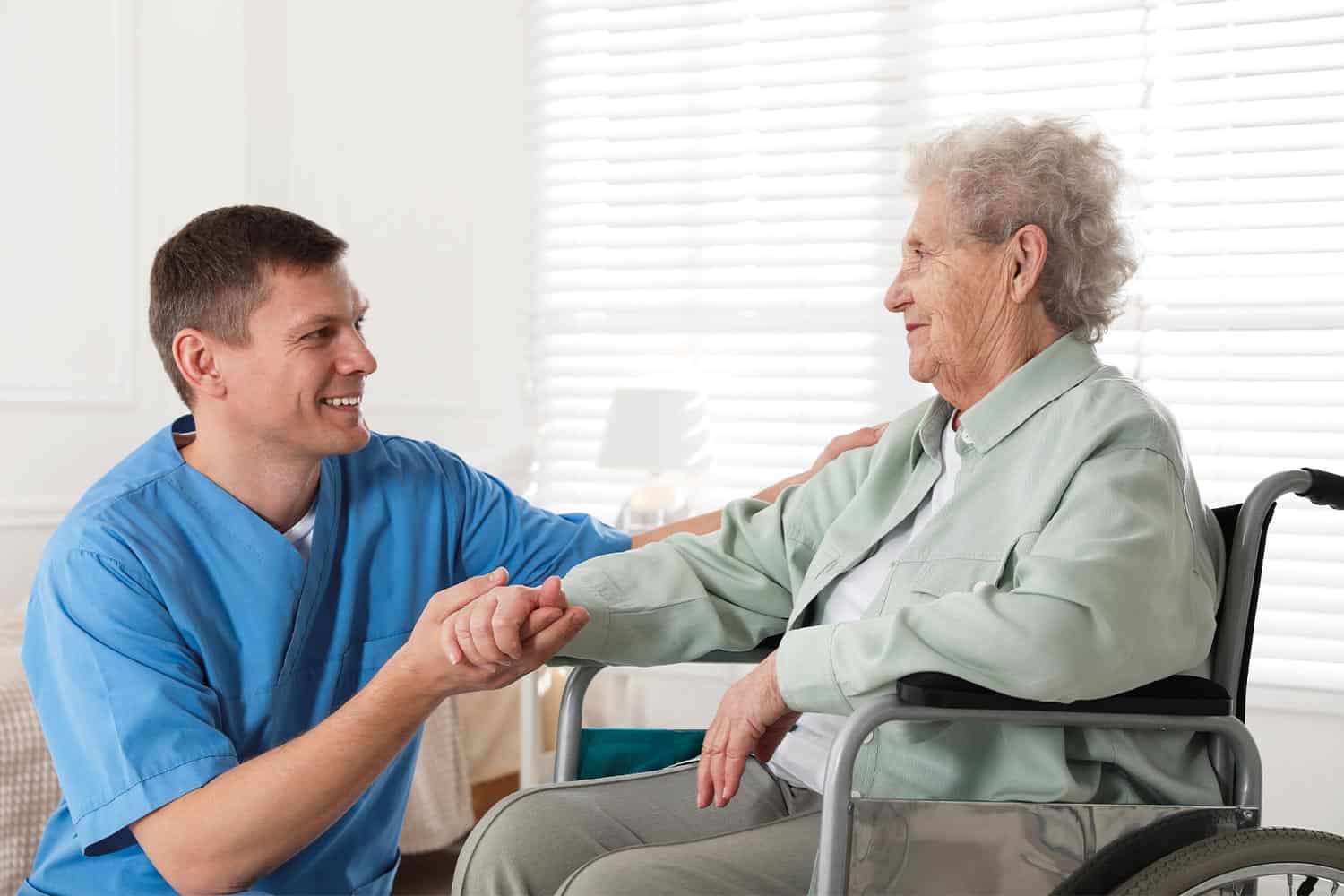 Male nurse sitting holding hand of elderly lady in wheelchair.