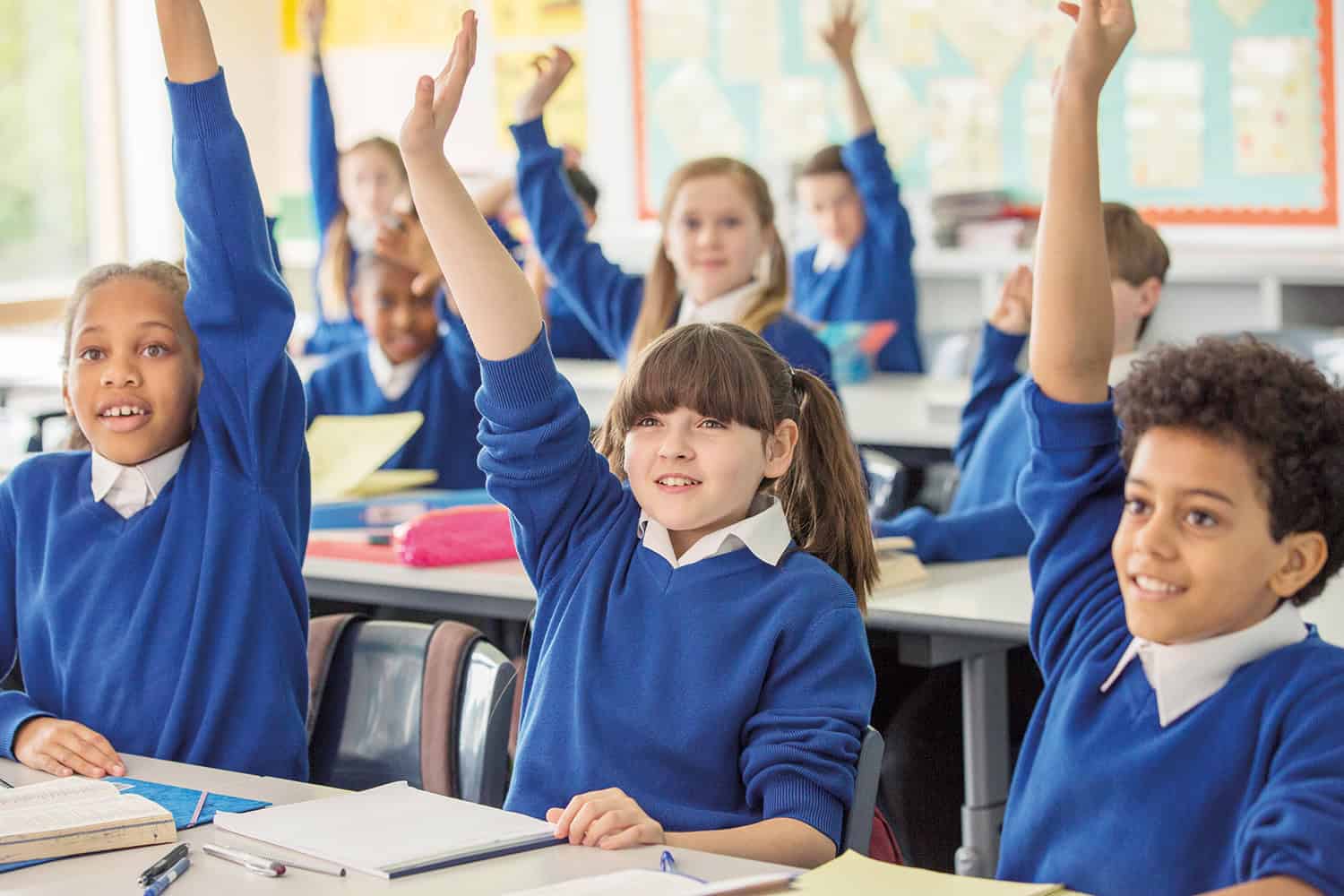 A classroom of pupils with their hands up in the air.