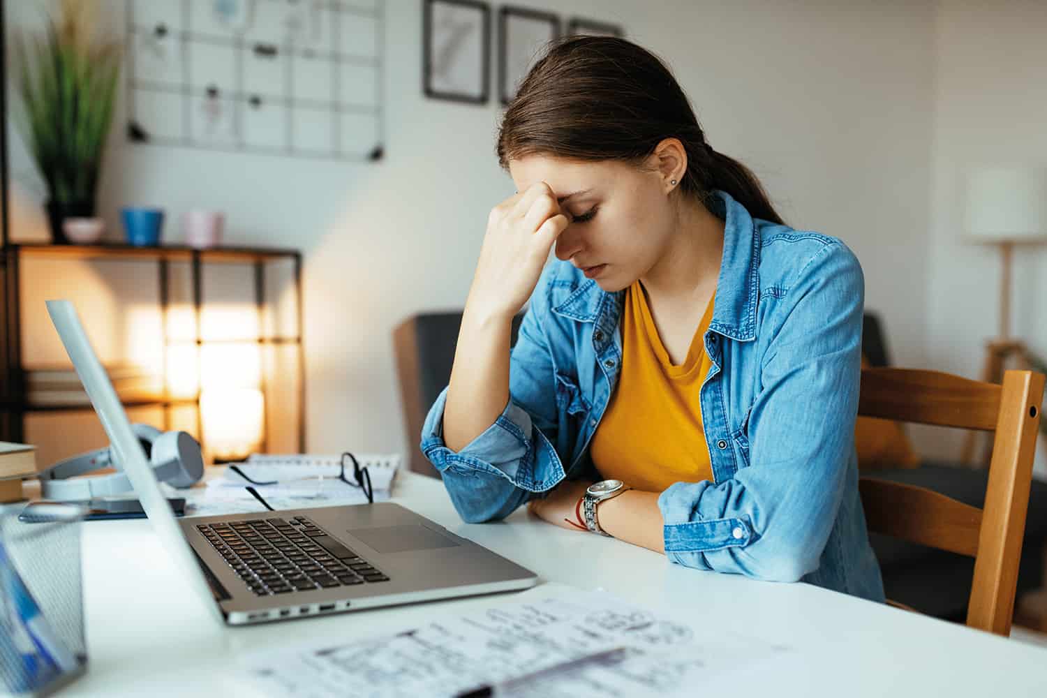 A woman sat in front of laptop, with hand on head, bowed.