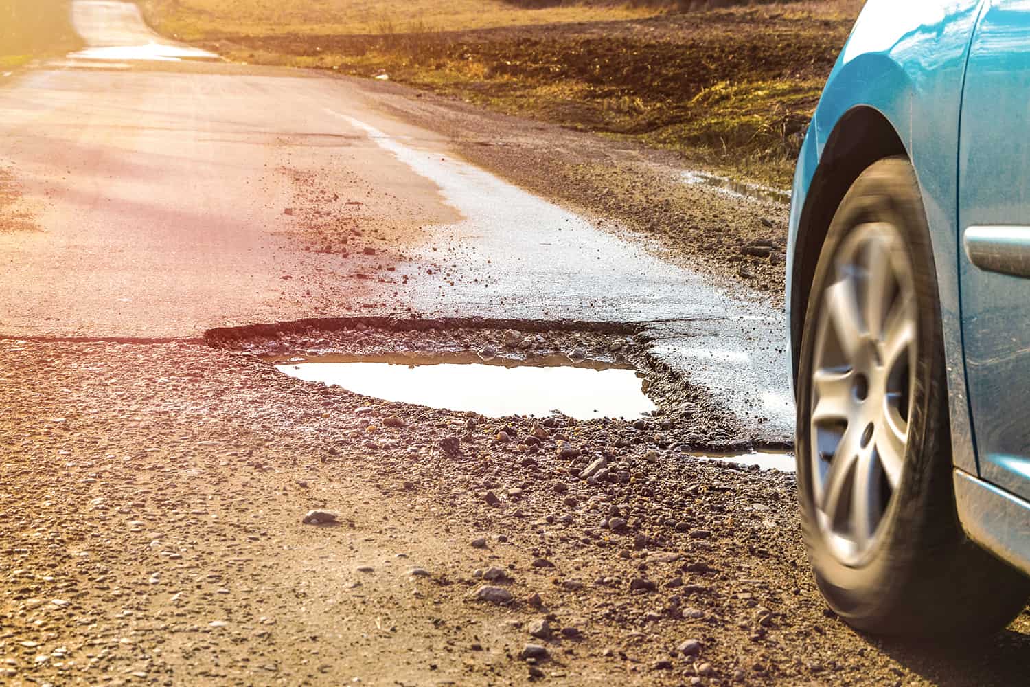 photo of close-up of front end of car about to go through a big, muddy, water-filled pothole on a narrow local rural road