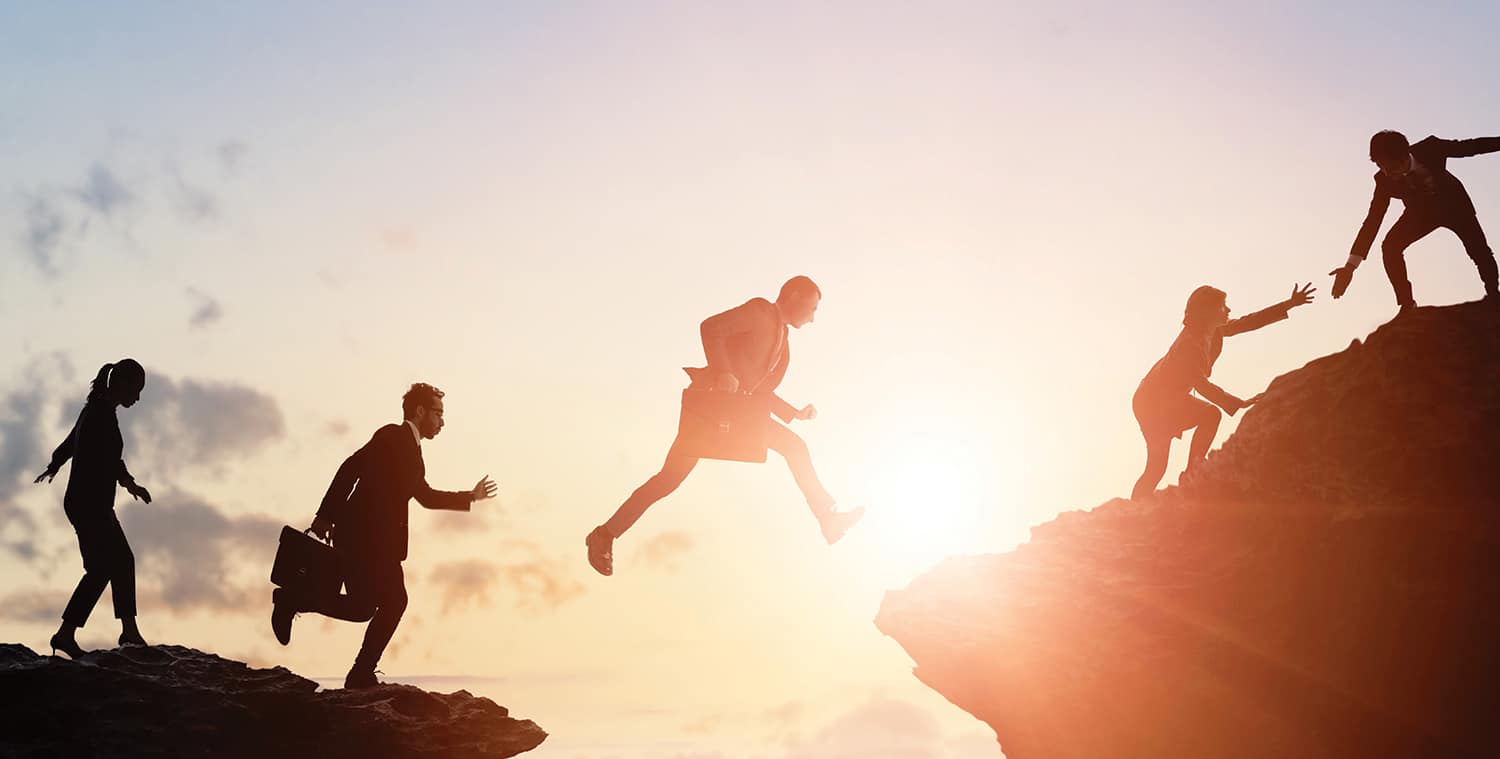 photo/graphic of businessmen and women helping each other across a cliff top and jumping across a gap, silhouetted against a rising or setting sun with mountains in the background