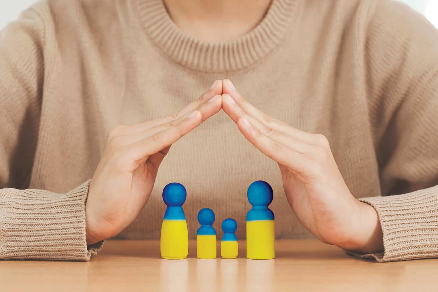 photo of hands forming a roof over a family of wooden peg-people painted in the blue and yellow of the Ukrainian flag