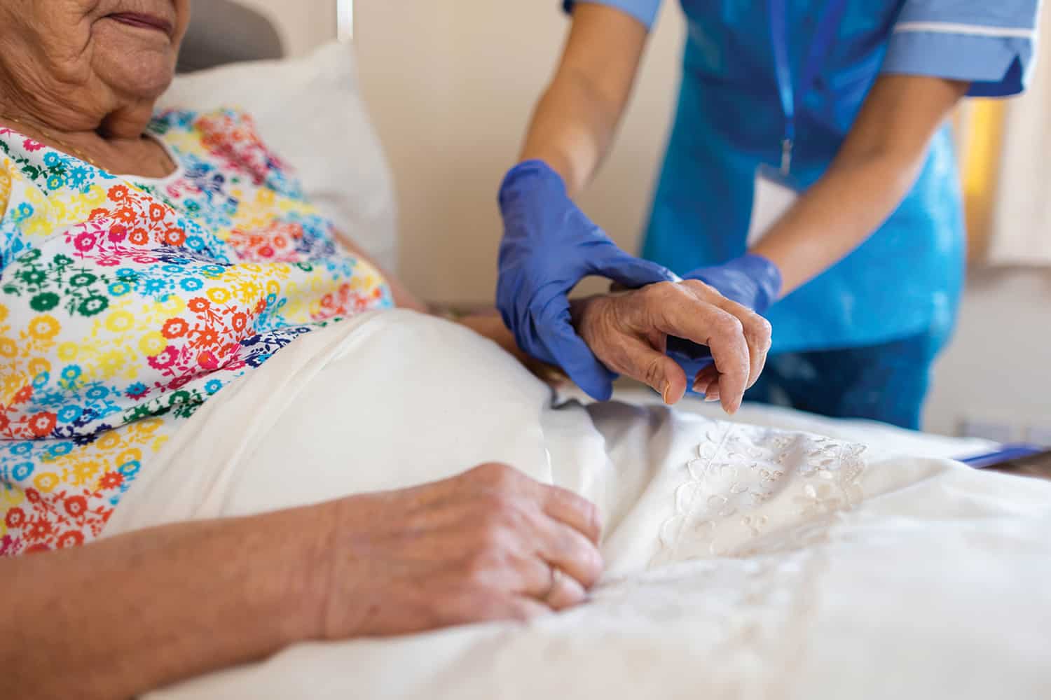 : care worker’s disposable-gloved hands holding wrist of older woman in bed wearing colourful night shirt