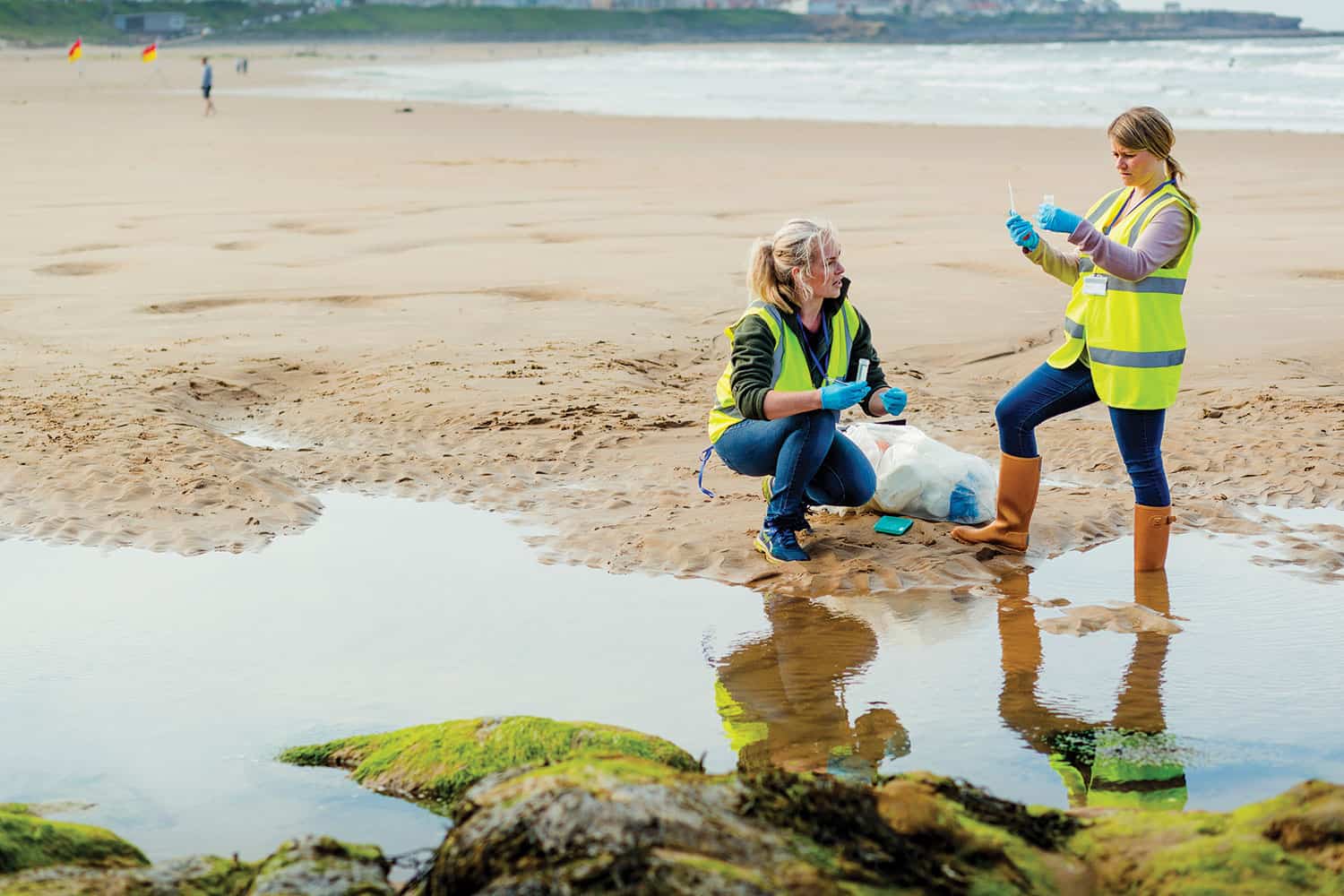 Pic: photo of two women in high-vis jackets and wellies sampling water in pool on beach with sea behind them.