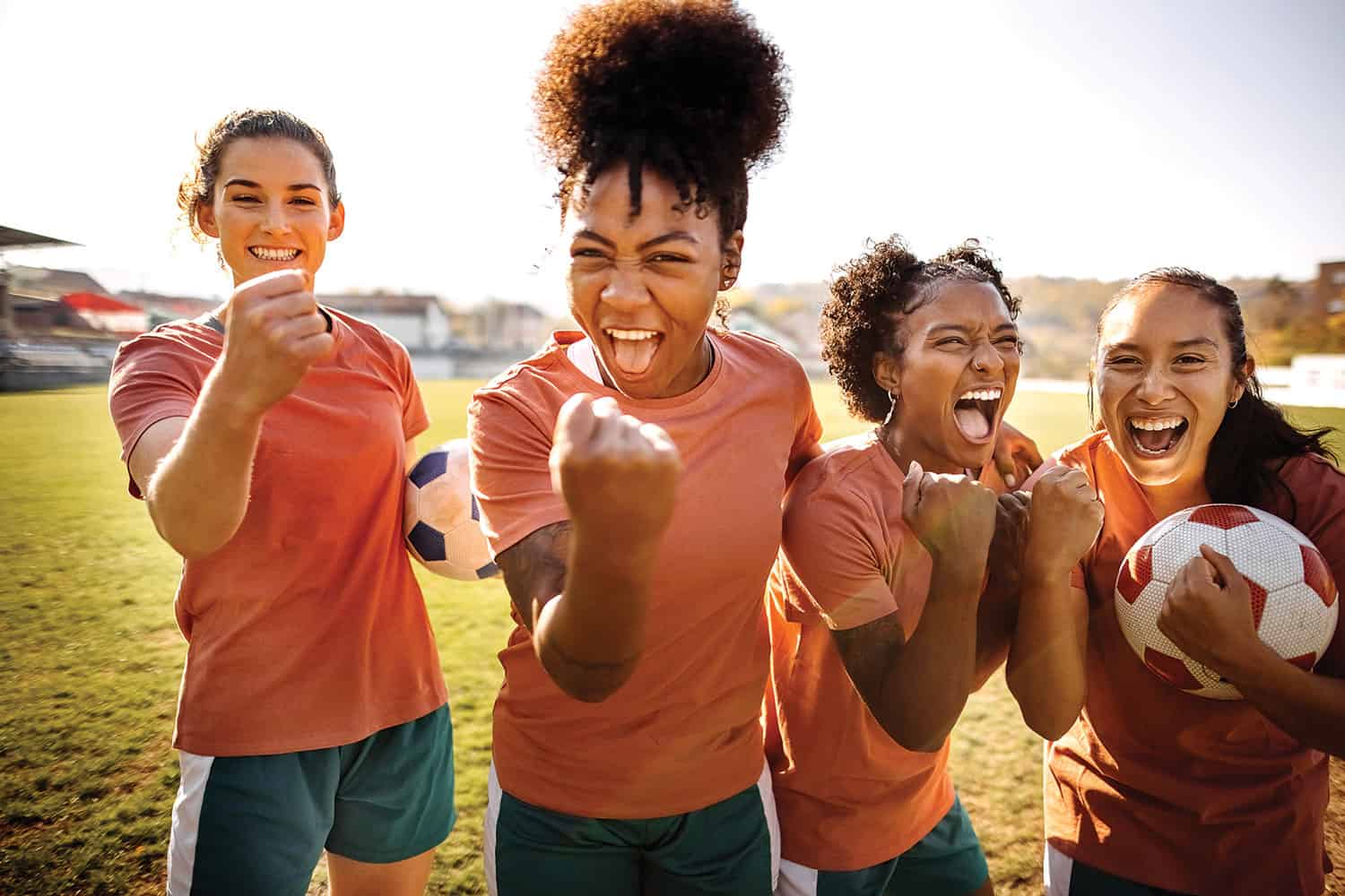 photo of four jubilant women footballers, smiling with fists raised, football pitch and stand in background
