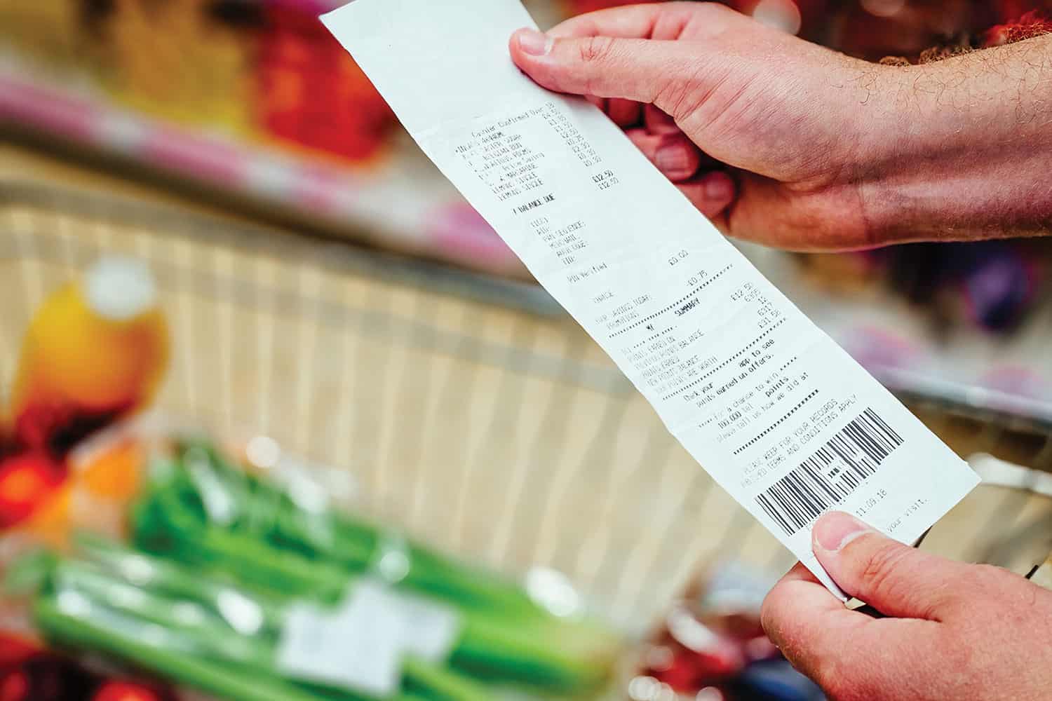 Photo close-up of hands holding supermarket receipt over shopping trolley containing groceries.