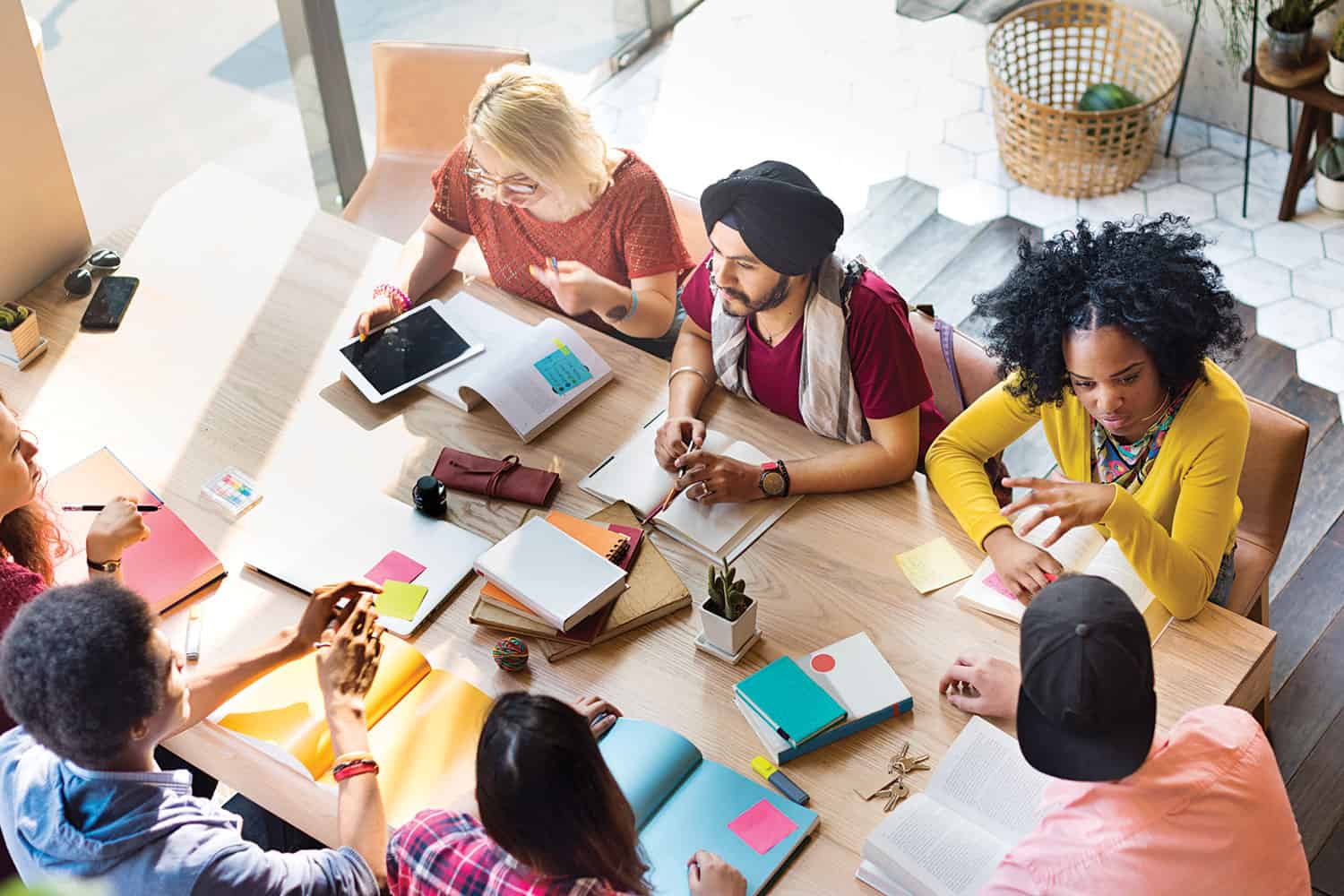 photo from above of mixed group of adult work colleagues sitting around a table scattered with notebooks, tablets and sticky notes in an office, discussing a project