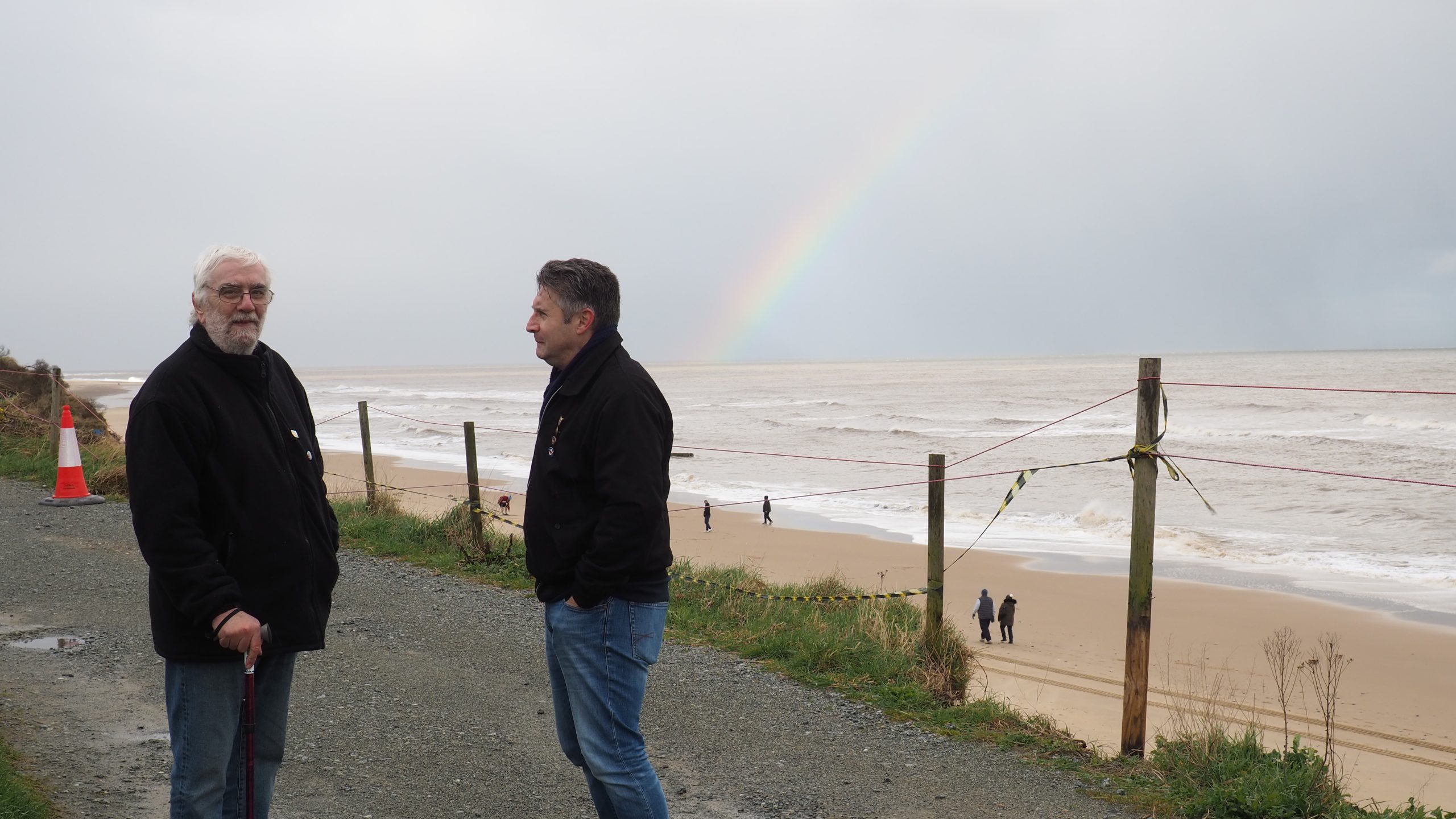 photo of two men, Kevin Jordan, left, and Simon Measures, members of Save Hemsby Coastline, standing on a gravel track above a beach with a rainbow over the sea, at Hemsby, Norfolk