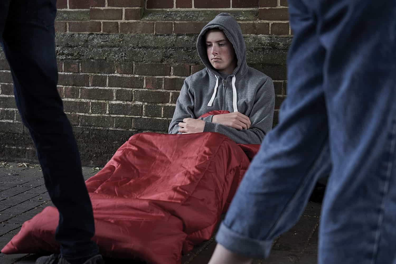 Young, sad homeless man in hoodie and red sleeping bag sitting on floor against brick wall behind legs of passersby.