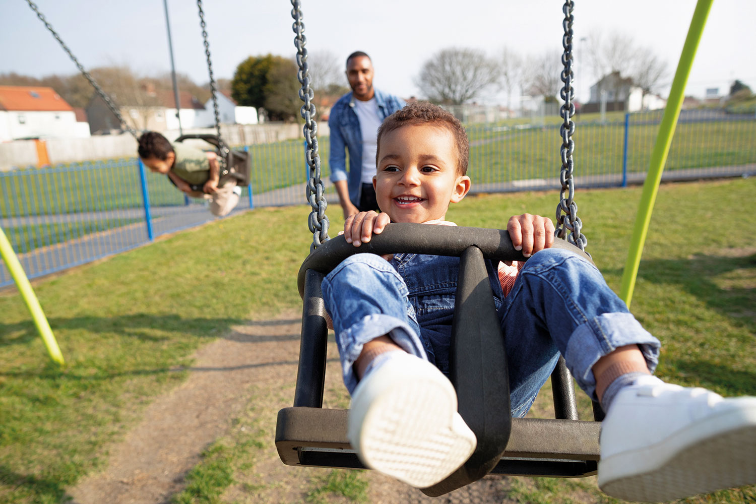 photo of smiling pre-school boy being pushed in a swing in an outdoor playground by his father who is in the background