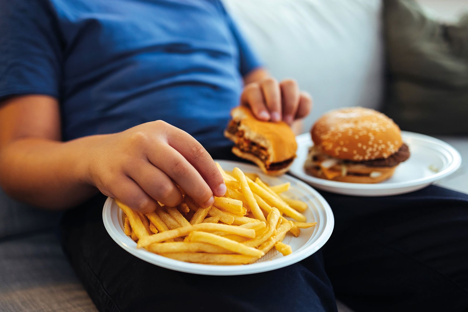 close-up of a boy’s hand picking chips off a plate, with a burger in his other hand