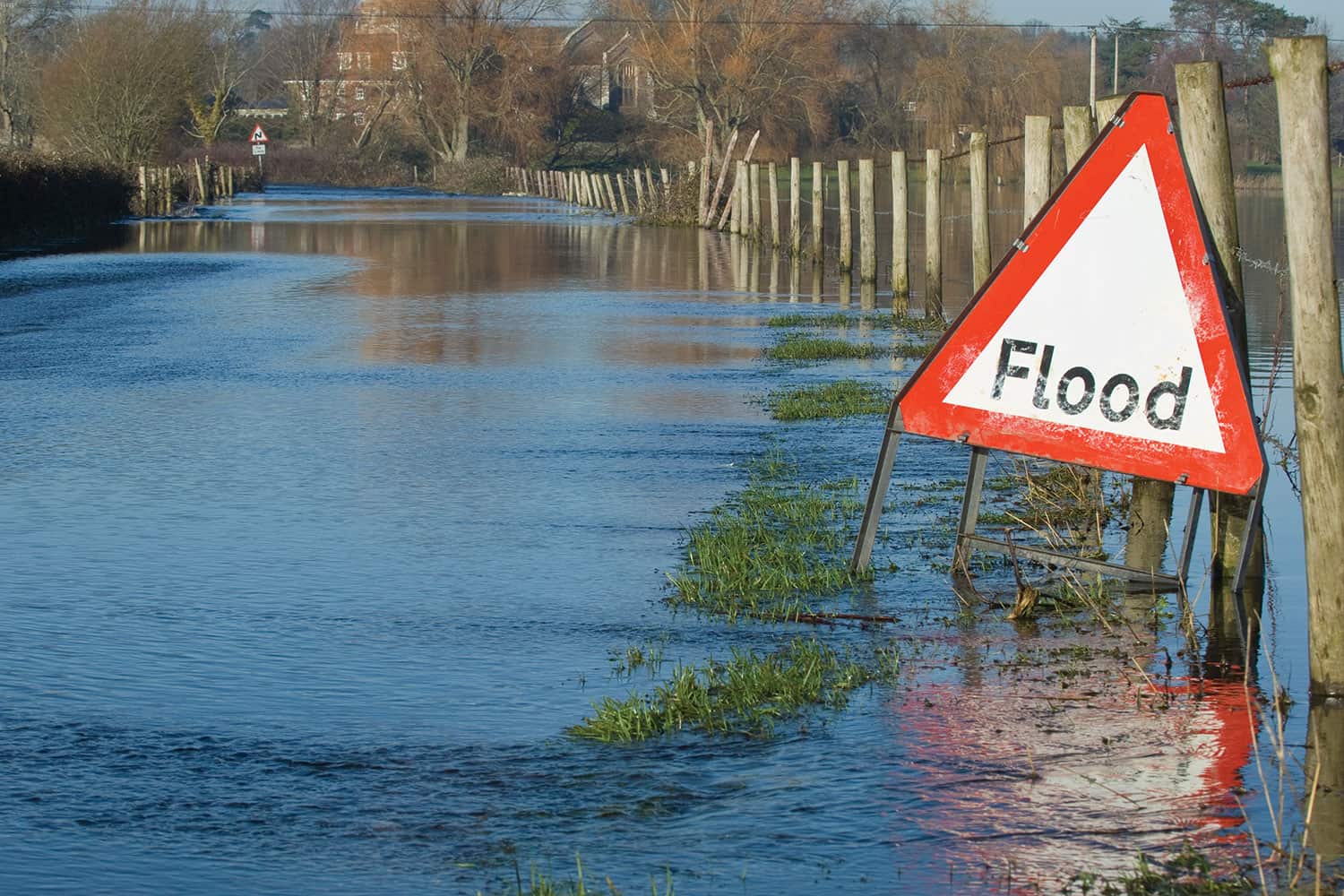 Red triangle ‘flood’ warning side set against flooded wooden fence with houses in background.