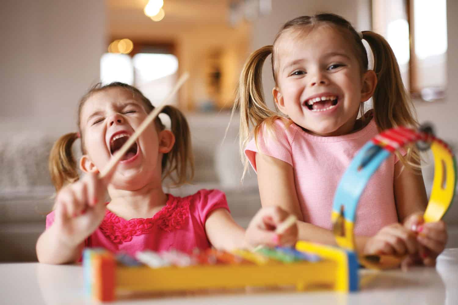 Two laughing, smiling girls with their hair in bunches, banging a drum and shaking a colourful tambourine).