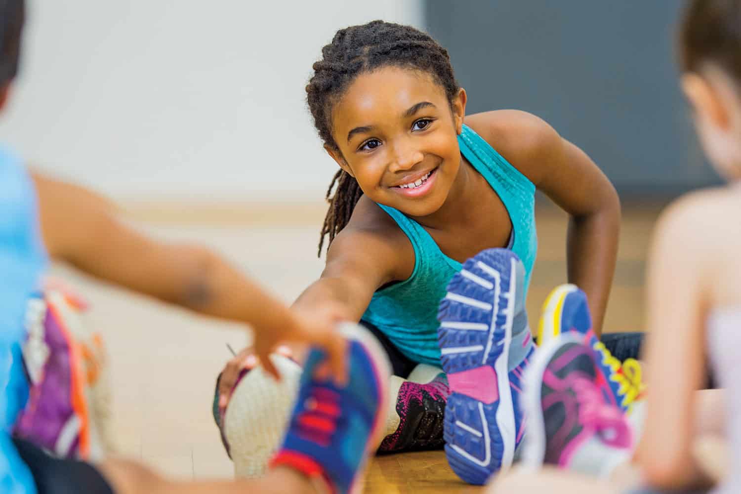 photo of smiling primary school-age girl sitting on a gym floor stretching to touch her toes, viewed between two other children who have their backs to us