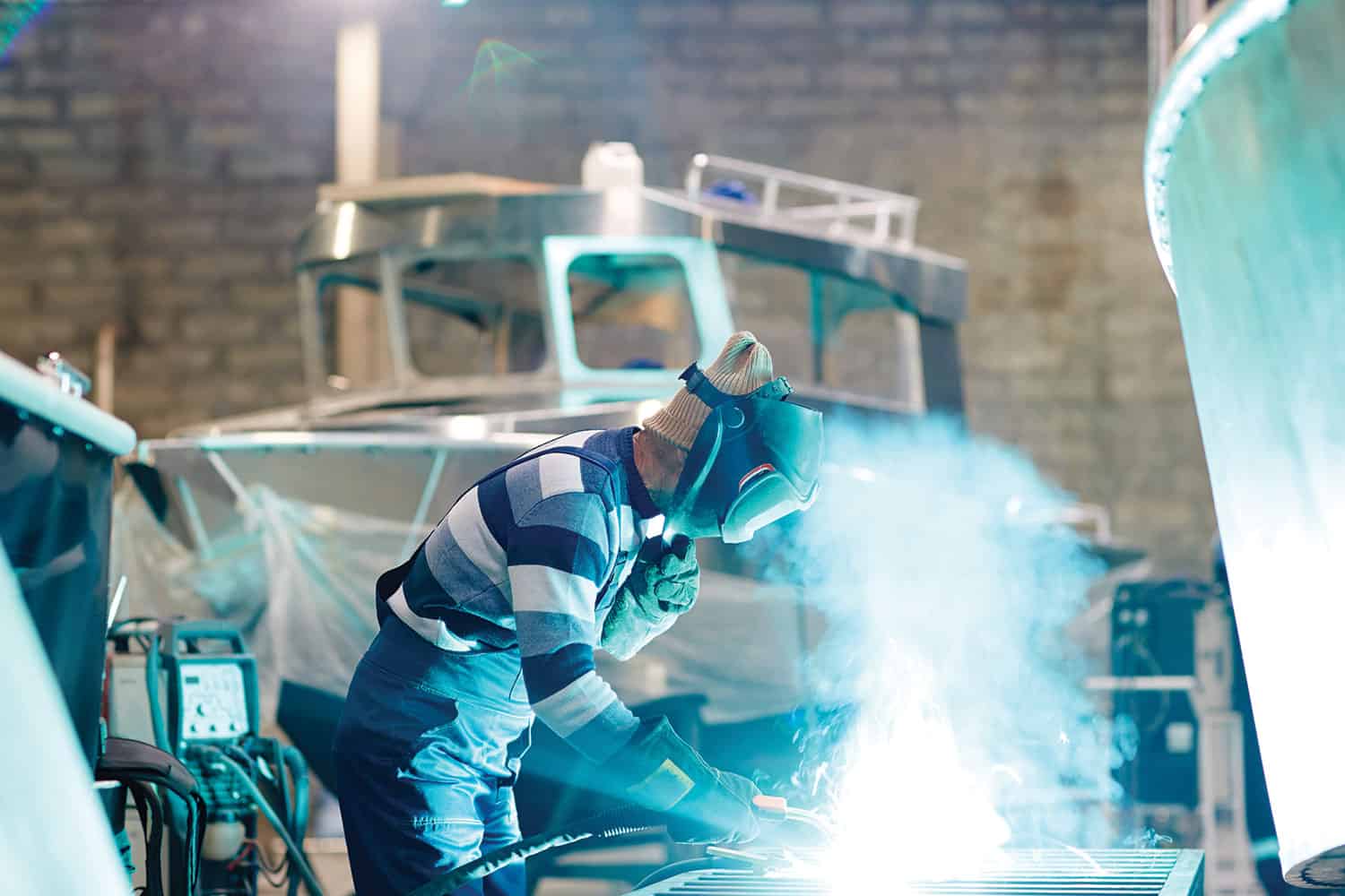 : photo of welder working with protective gloves and mask in front of a partly-built boat