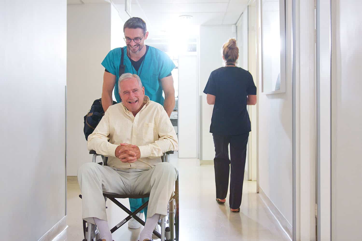 photo of older man being wheeled through a hospital corridor by a smiling young porter, with the older man’s bag on his shoulder