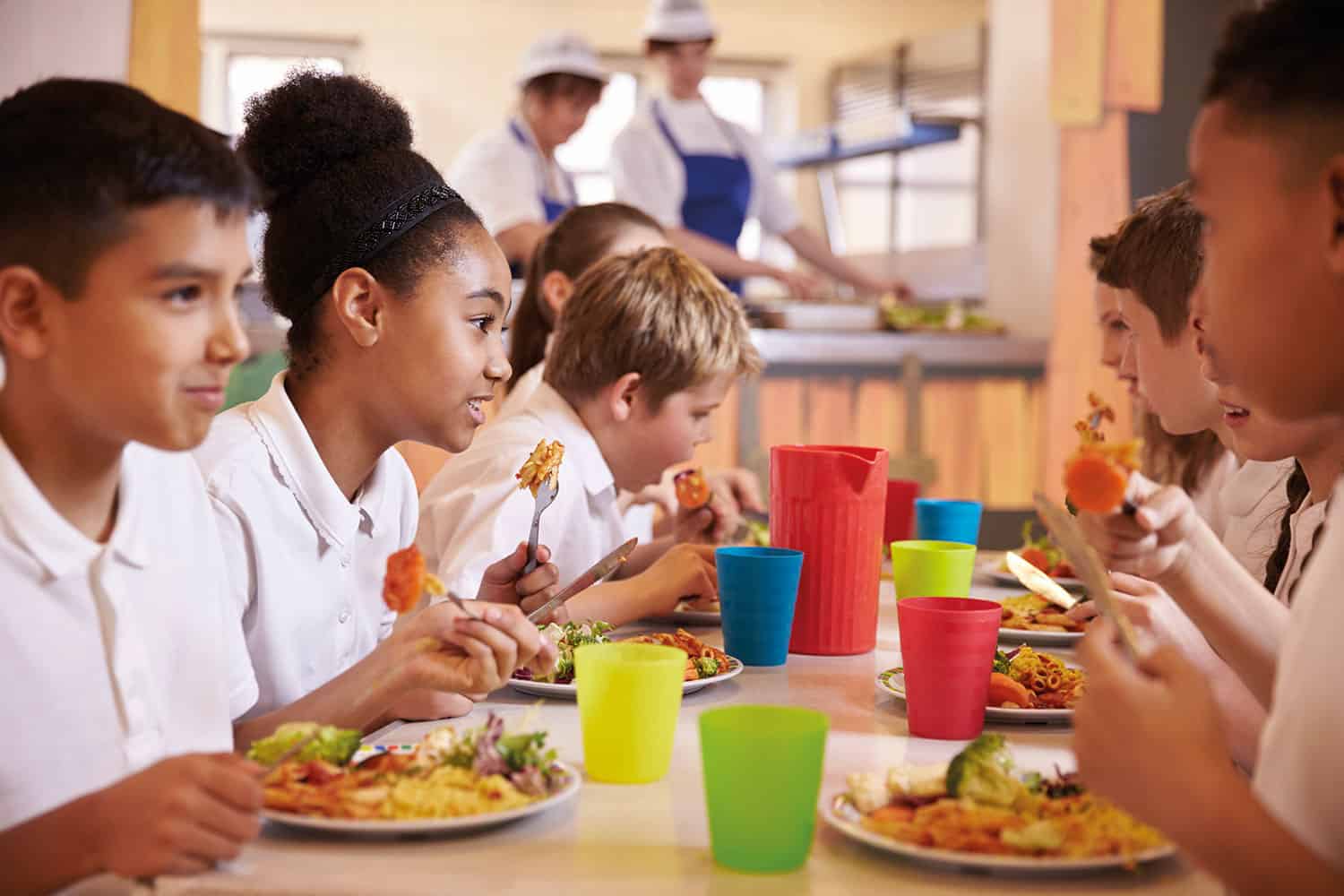 Photo of primary-age school pupils at table eating school dinner, amid colourful beakers and water jugs.