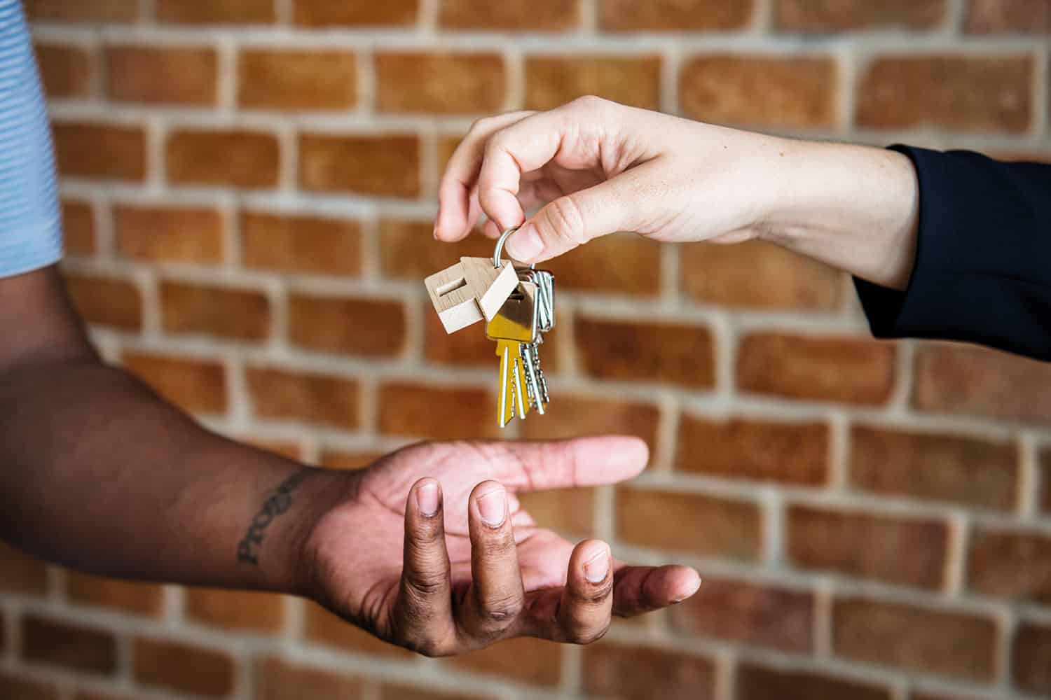 photo close-up of one person’s hands dropping house keys into another’s hands, against a backdrop of a red-brick wall).
