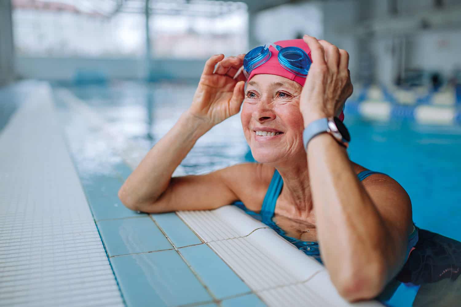 photo of older woman in swimming cap and goggles on her forehead resting at the edge of a swimming pool and smiling