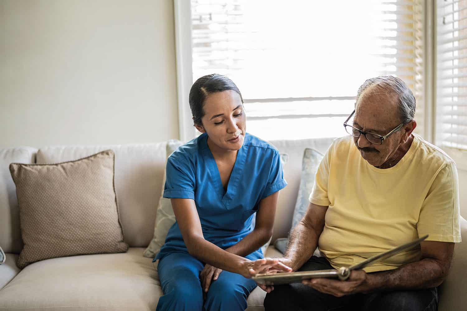 female carer in blue uniform sitting on sofa with older man, looking at tablet
