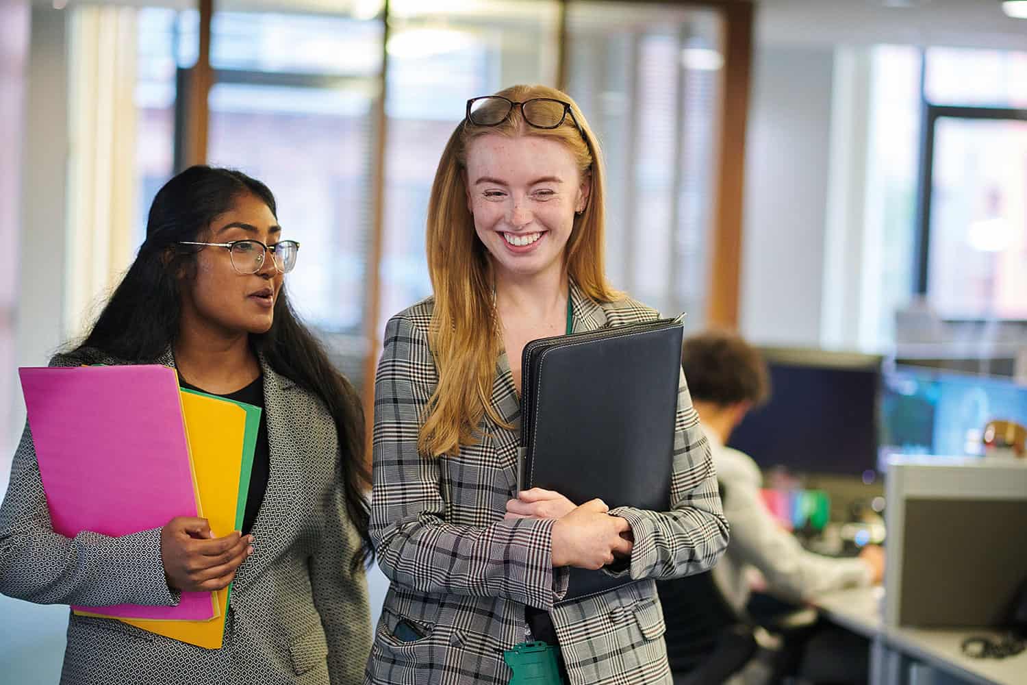 two young women carrying folders and chatting as they walk through an office