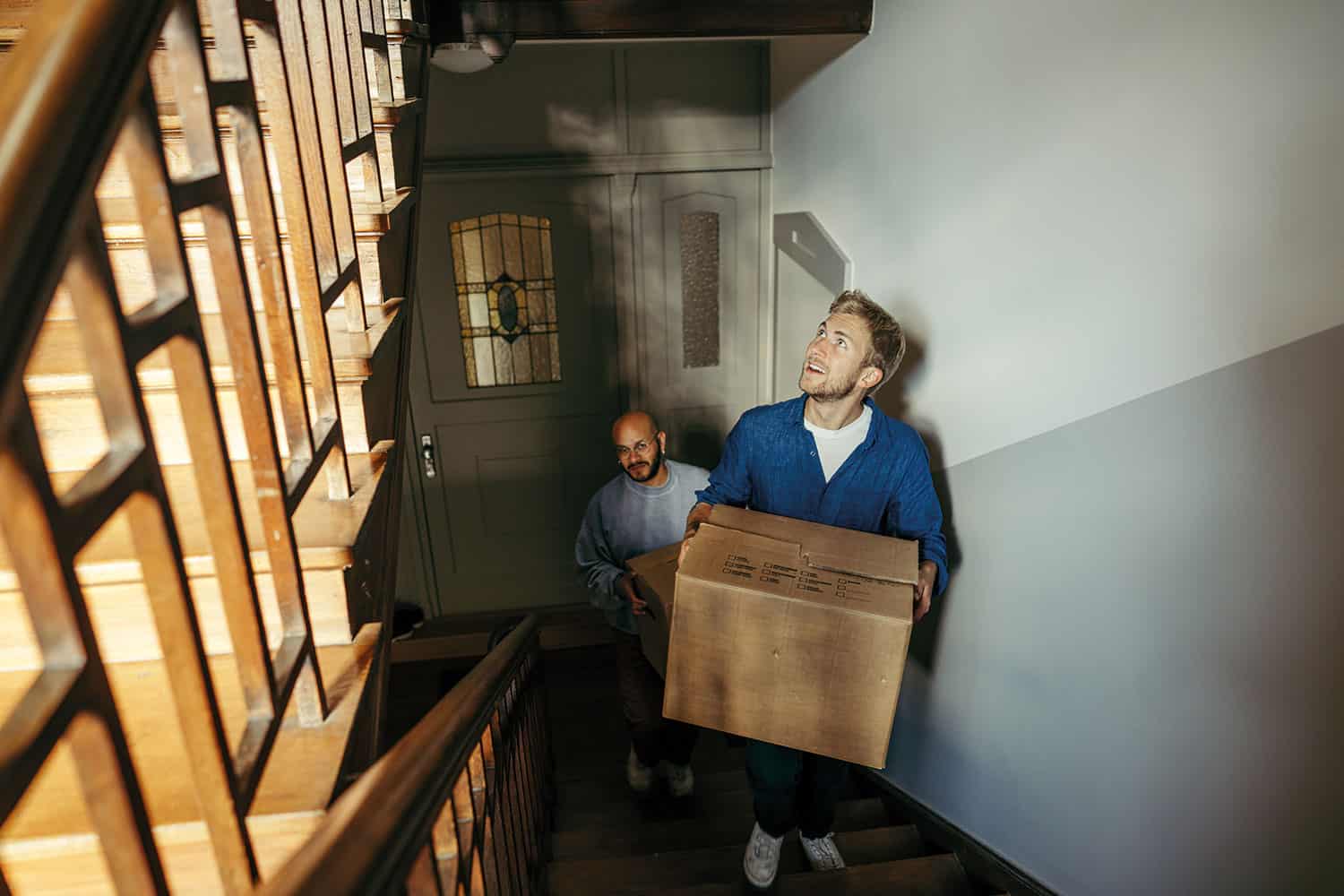 photo of young man carrying box up stairs, hall and front door behind him