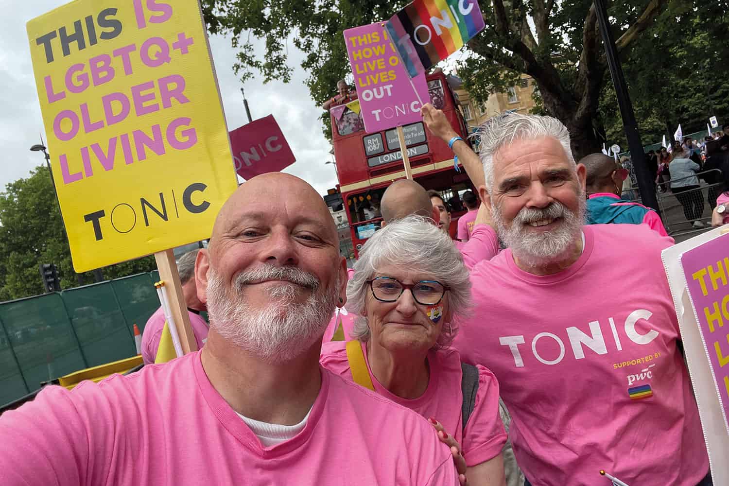 photo of Terry Stacy, Chair of Tonic Housing, with supporters Lorraine Shears and Paul Symes in pink t-shirts and amid colourful placards at London Pride 2025