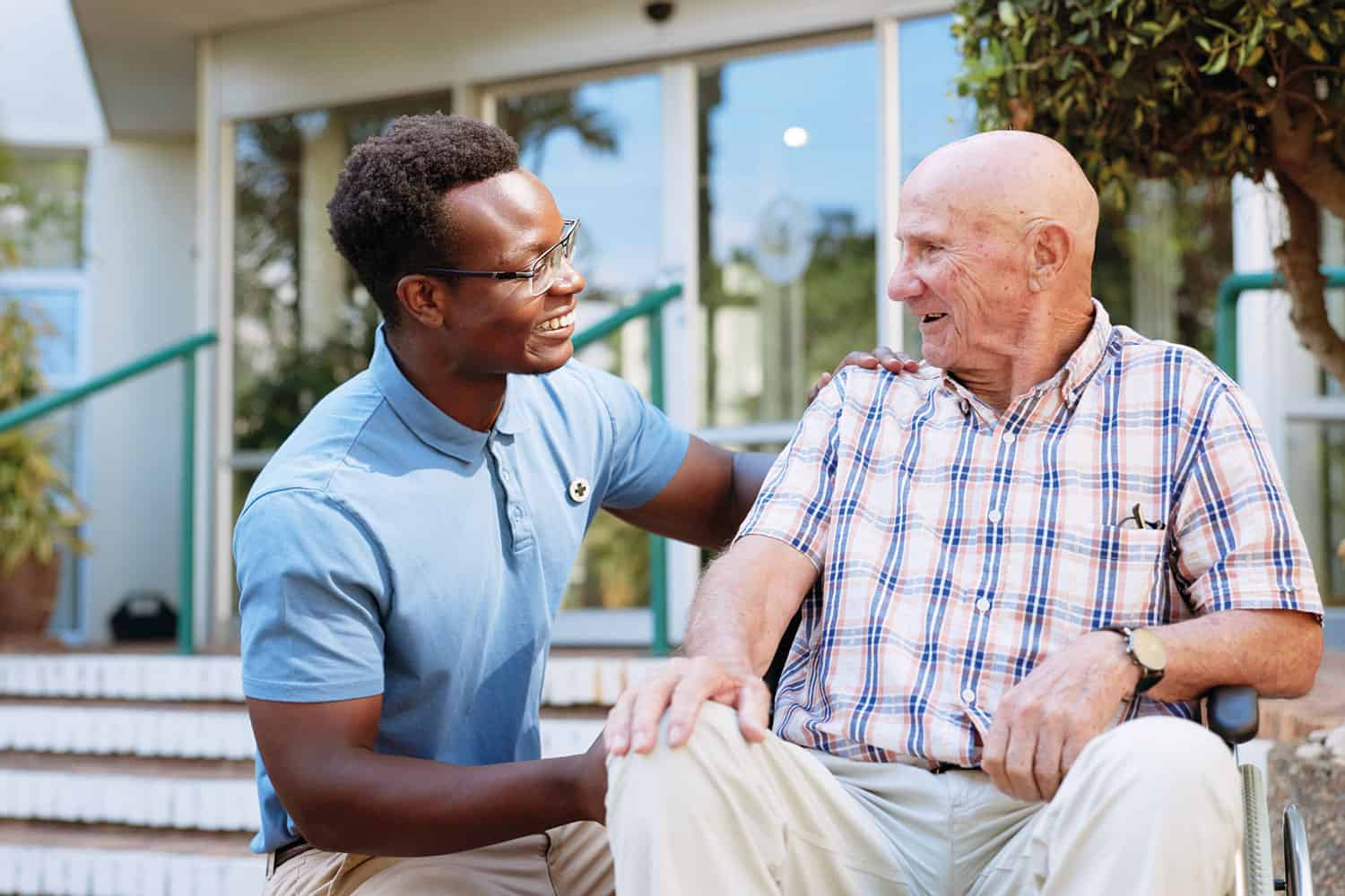 photo of older man sitting in chair outside home in sunshine, with younger male carer kneeling beside him