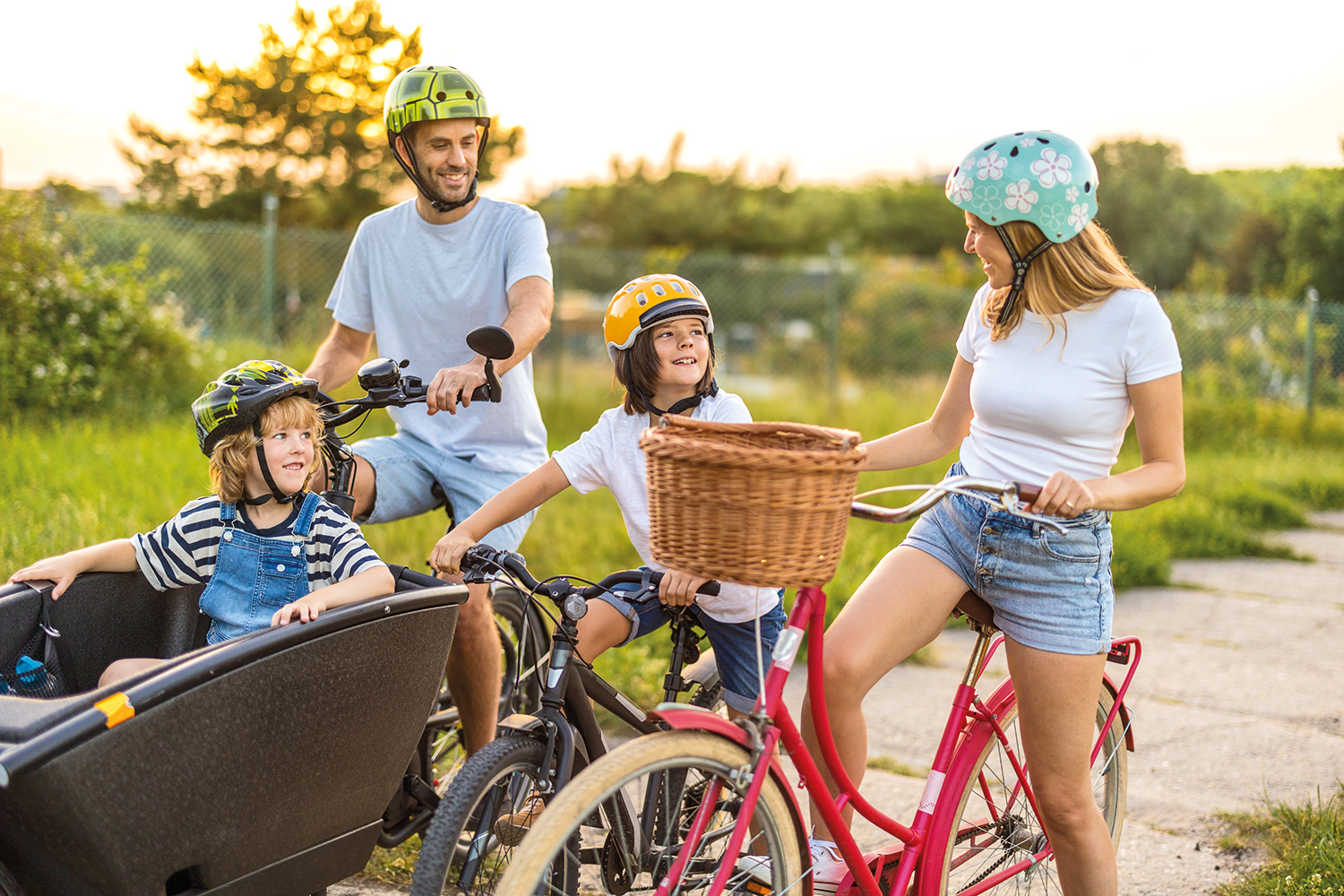 mum, dad and older kid on push bikes, with a younger kid riding in a cargo box at the front of the dad’s bike, all in t-shirts, shorts and helmets on a sunny day