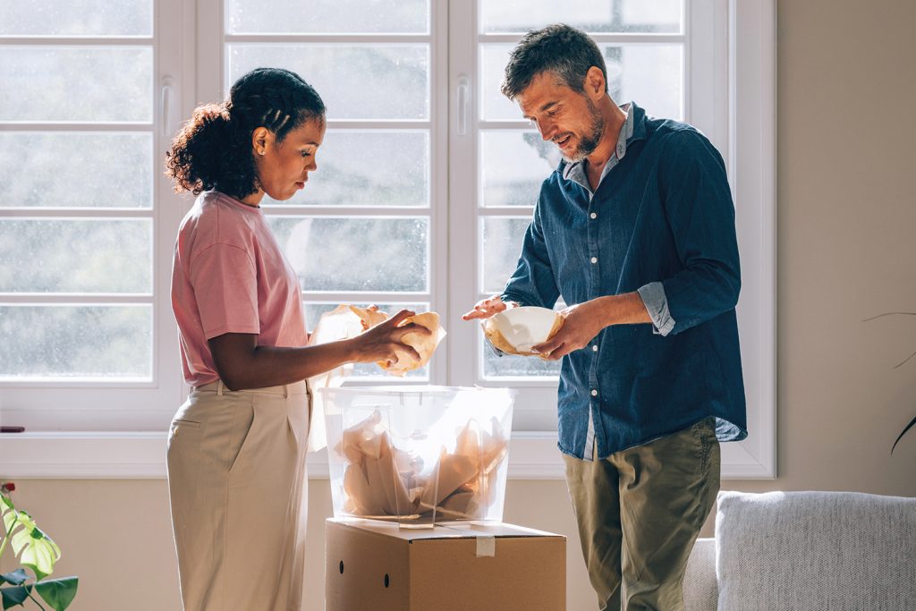 couple unpacking crockery from a moving box in a brightly lit front room full of boxes