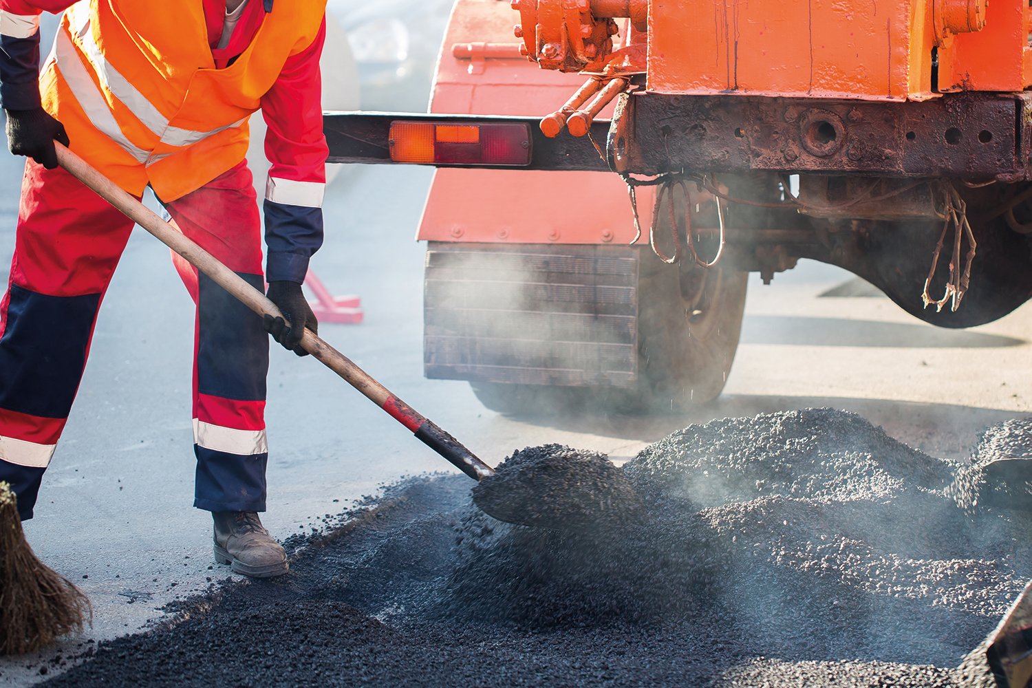 worker in high-vis orange uniform shovelling tarmac stones behind a road-laying machine
