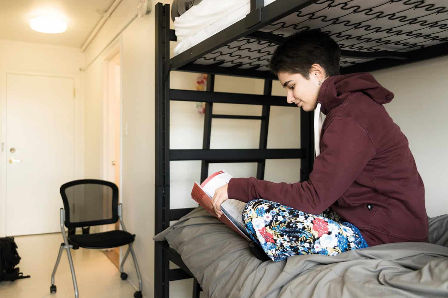 photo of boy aged 11-14 sitting cross-legged reading a book on the bottom bed of a bunk bed in a bare room
