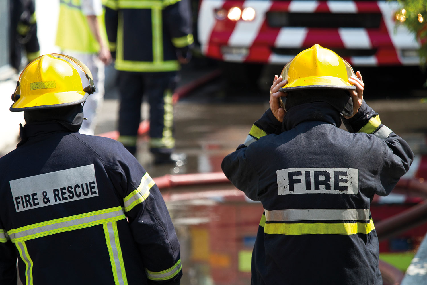 photo from behind of two firefighters in full operational gear with yellow helmets, with fire engine and hose and water in the background