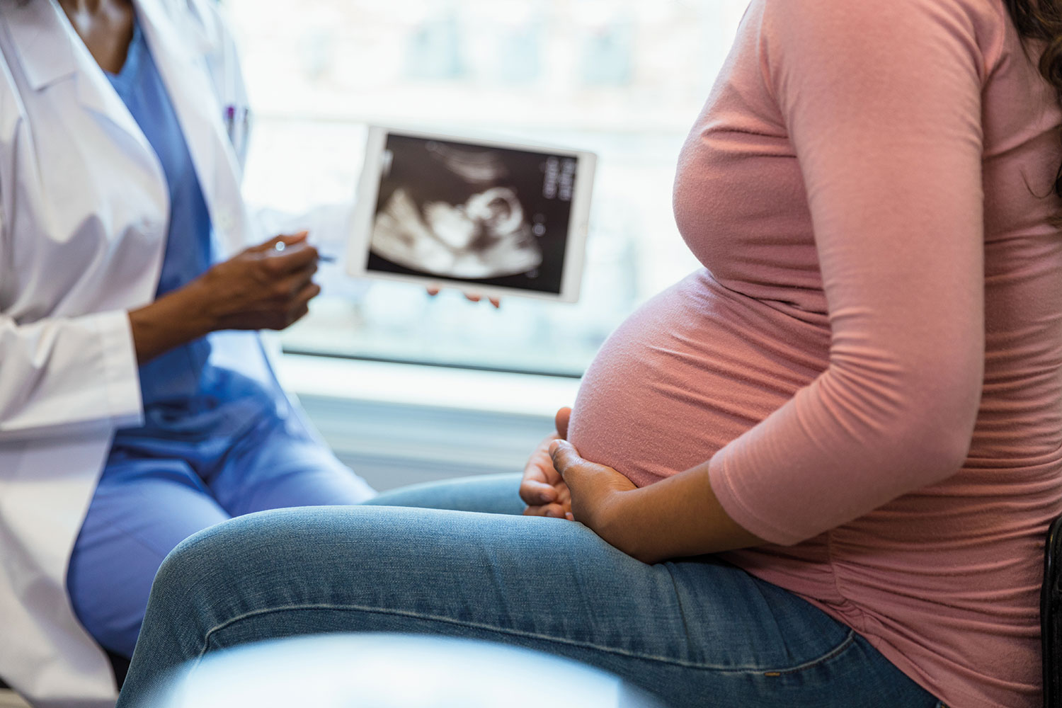 photo focused on mid-sections of a medic in a white coat talking to a heavily pregnant woman in a pink top, with a baby scan on a screen between the two of them