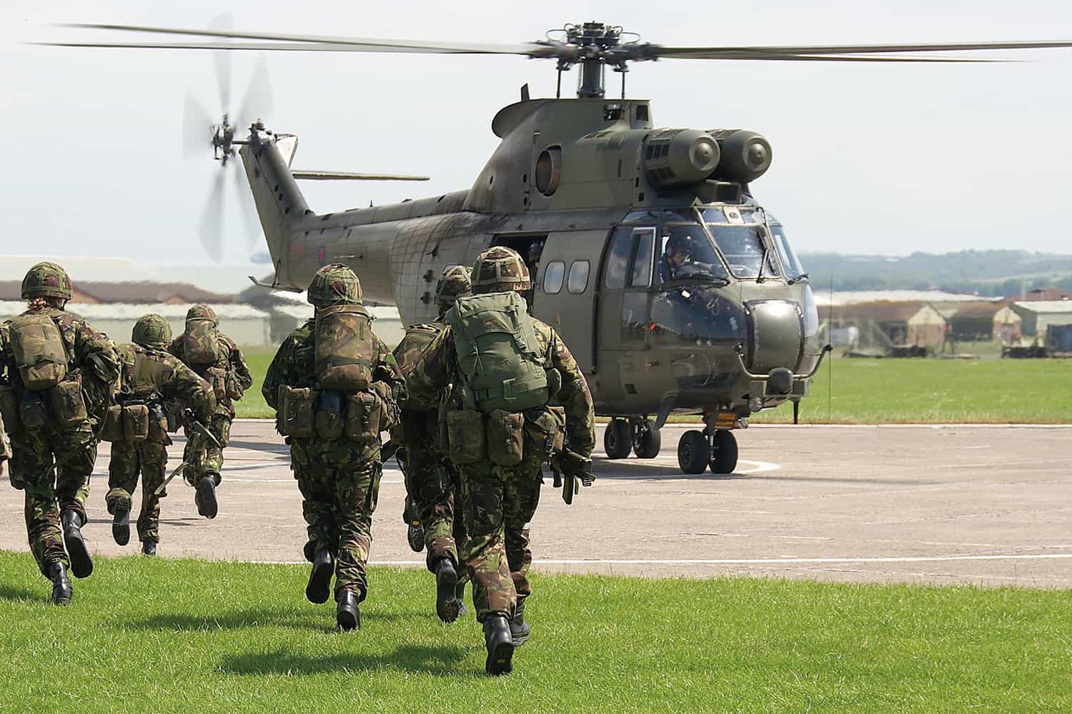 photo of six soldiers from behind in full combat greens, helmets and rucksacks, running towards a military helicopter with rotors spinning parked on concrete circle in green field with military camp in background