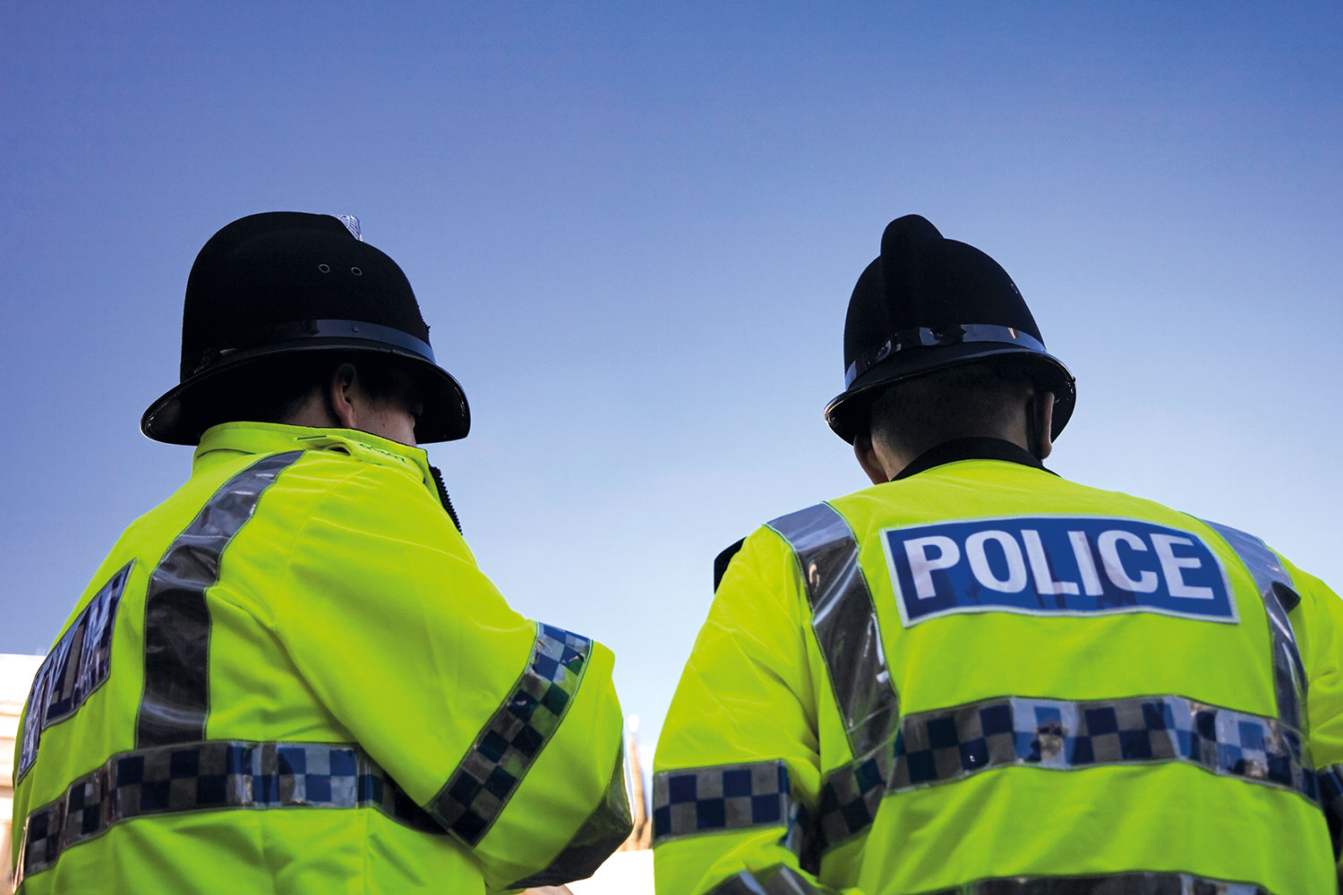 photo from behind and looking up at top-halves of two male police officers in high-vis jackets and helmets