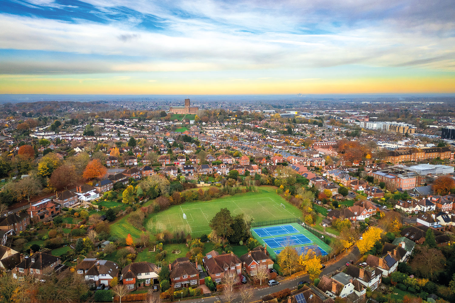 aerial photo of town, centred on a large playing field with astrocourts surrounded by housing stretching into a distant horizon of sunrise or sunset