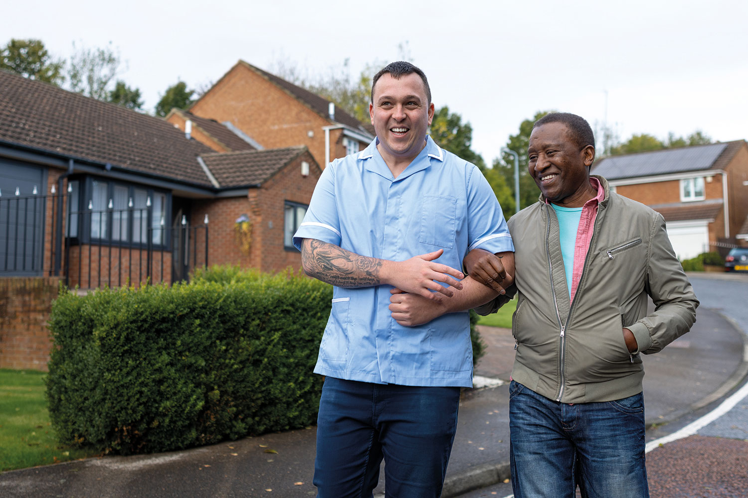 photo of older adult male with carer in blue uniform walking outside houses in north of England