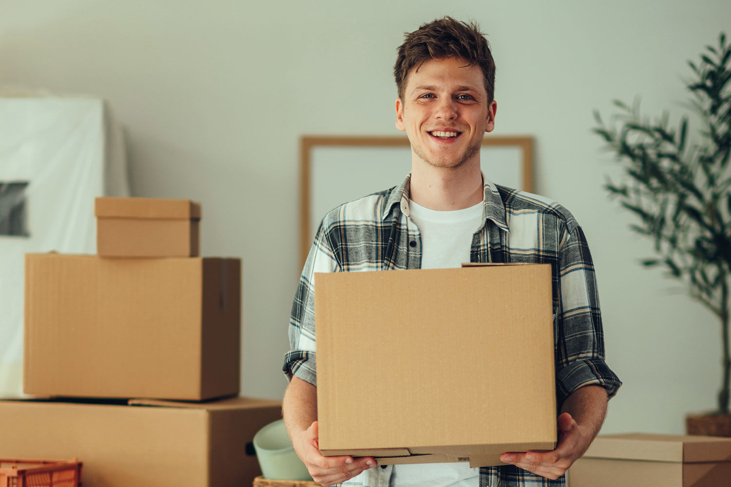 photo of young man smiling into camera, holding cardboard box with other boxes piled on tables behind him