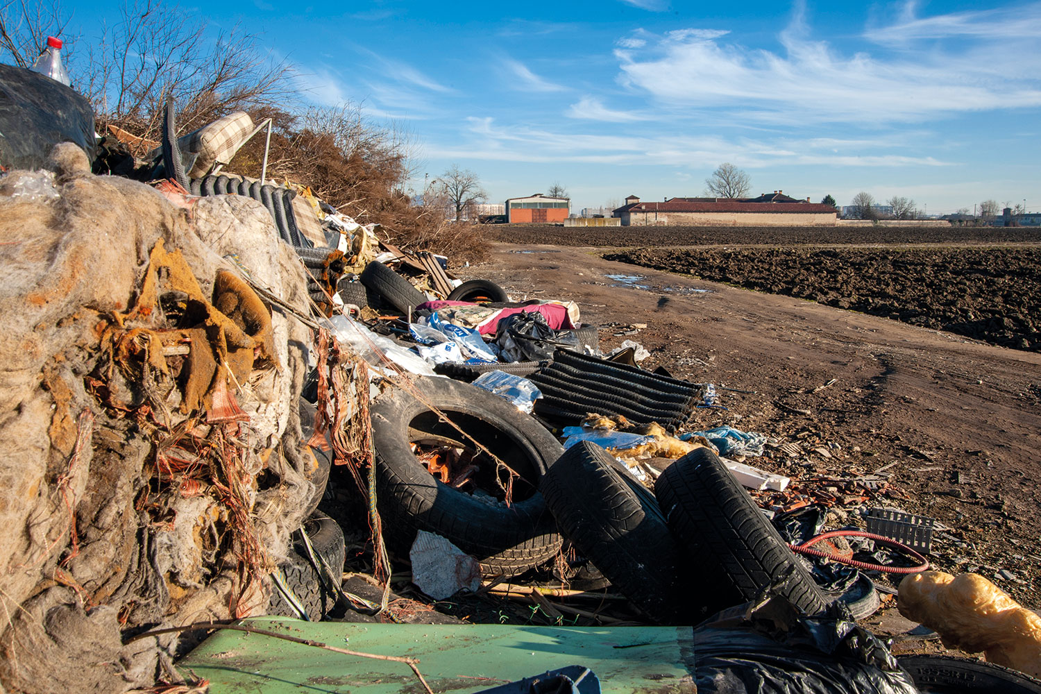 close-up of fly-tipping heaped up against an embankment, including tyres, shoes mattresses and industrial waste, at the edge of a ploughed field with farm buildings in the background