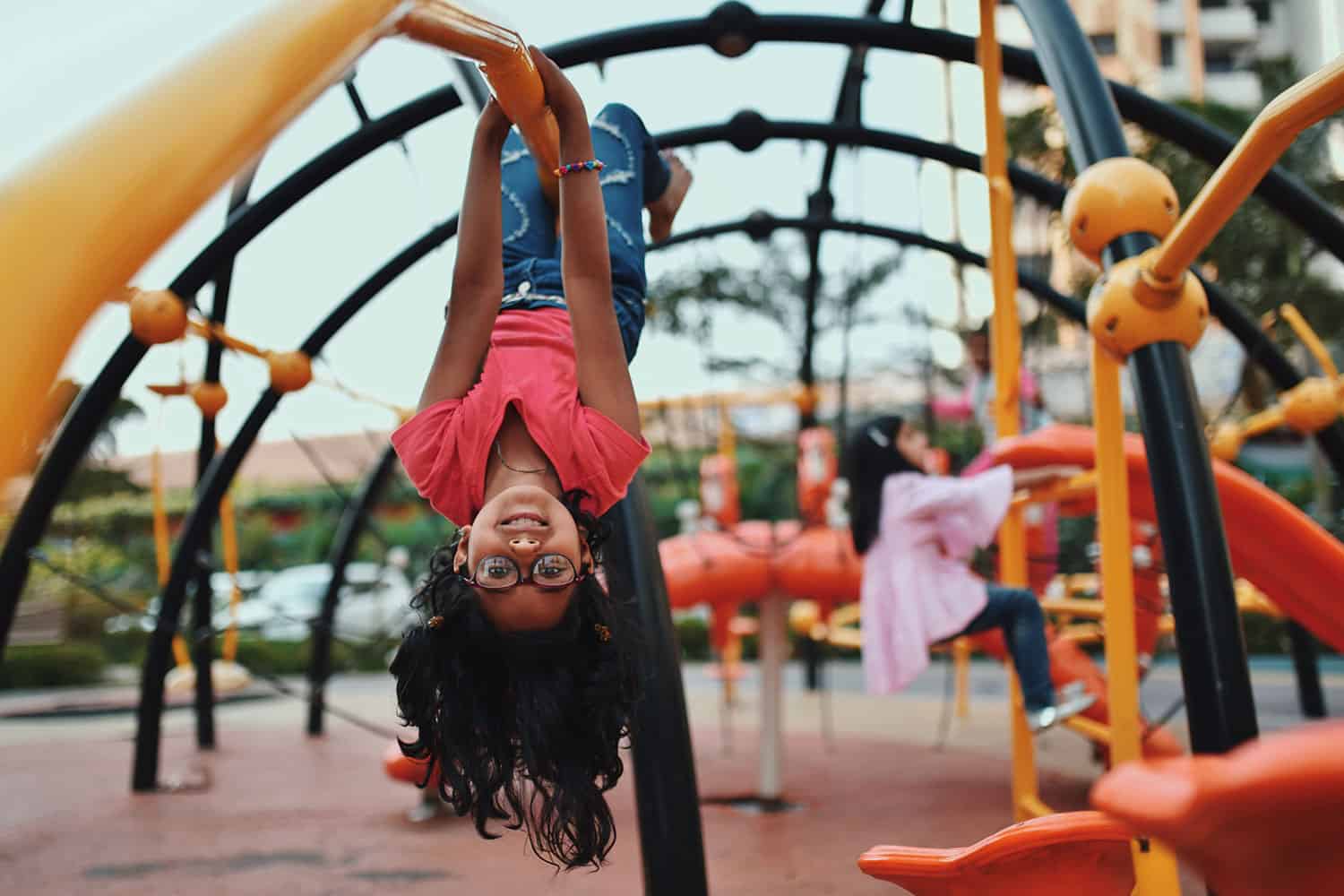 photo of young girl hanging upside down on colourful climbing equipment in a playground with another child in the background
