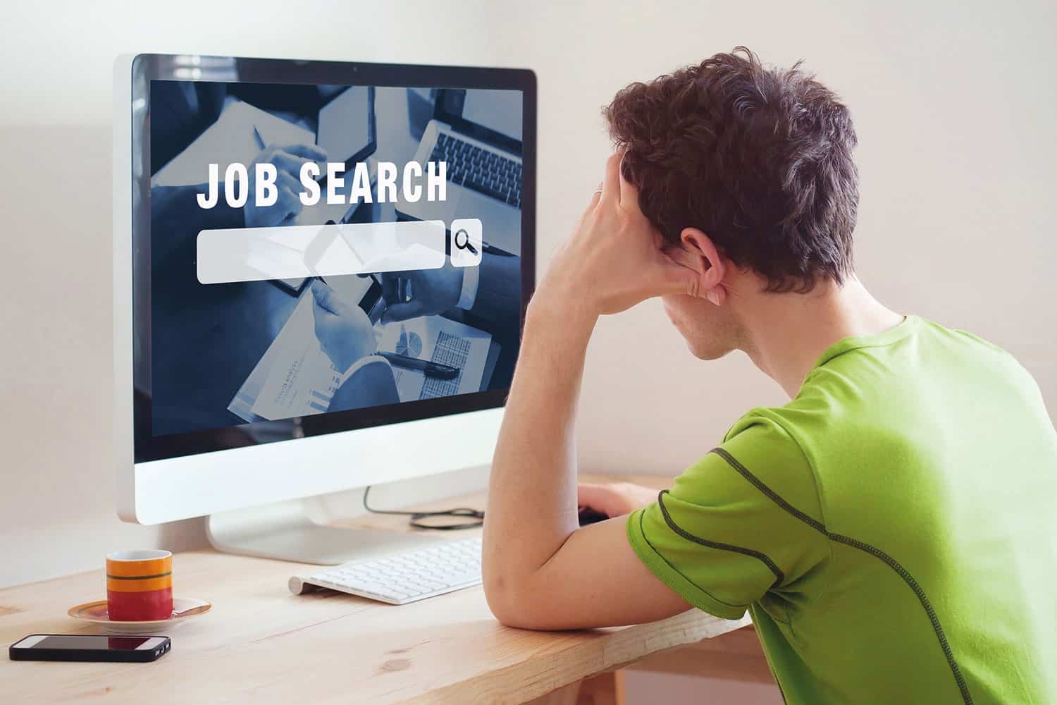photo from behind of young man in green t-shirt sitting at a desk looking at a computer screen with ‘JOB SEARCH’ in big letters on it, with his head in one hand