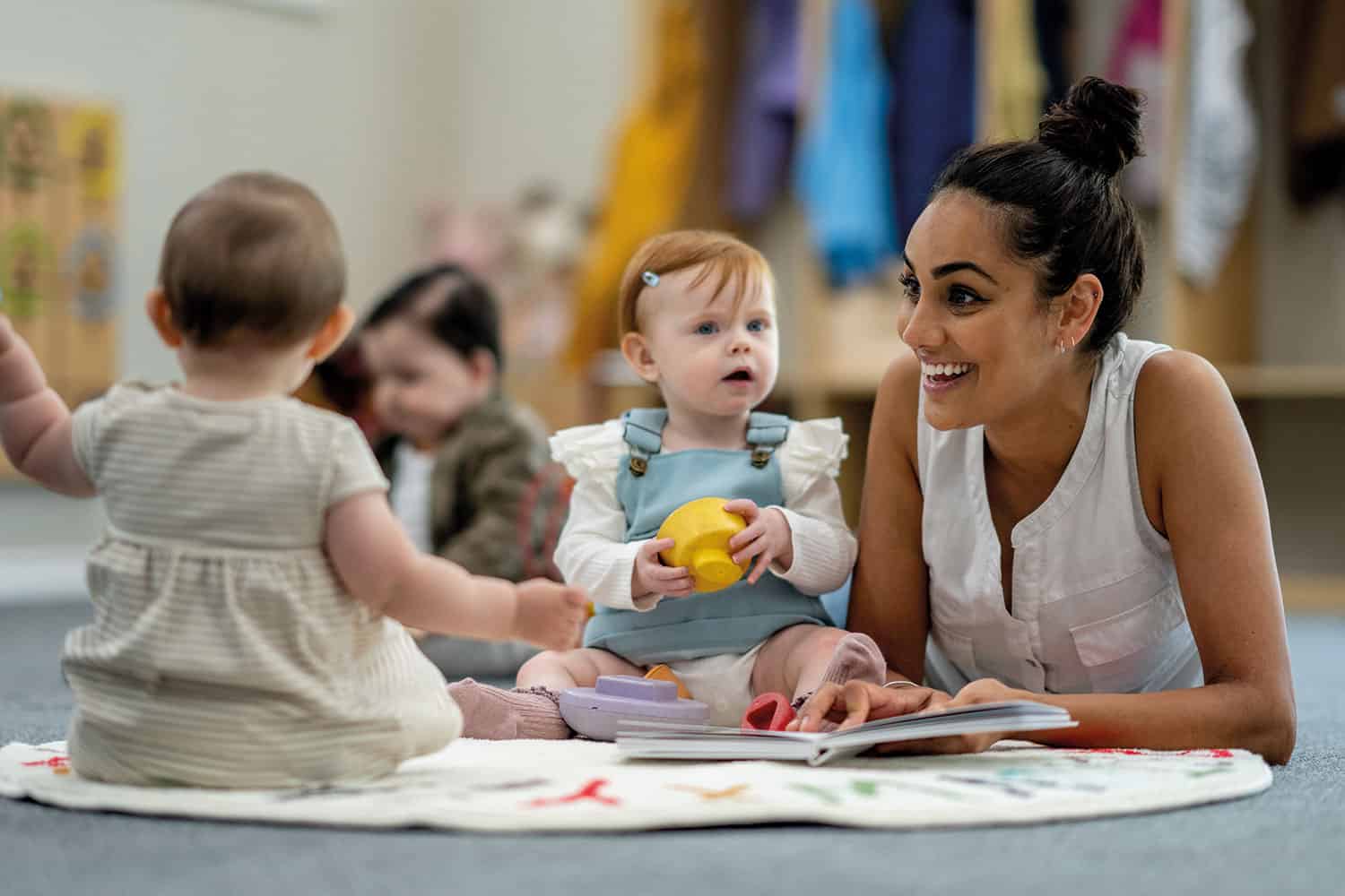 photo of young female nursery worker on floor with two babies, a book and shaking toys