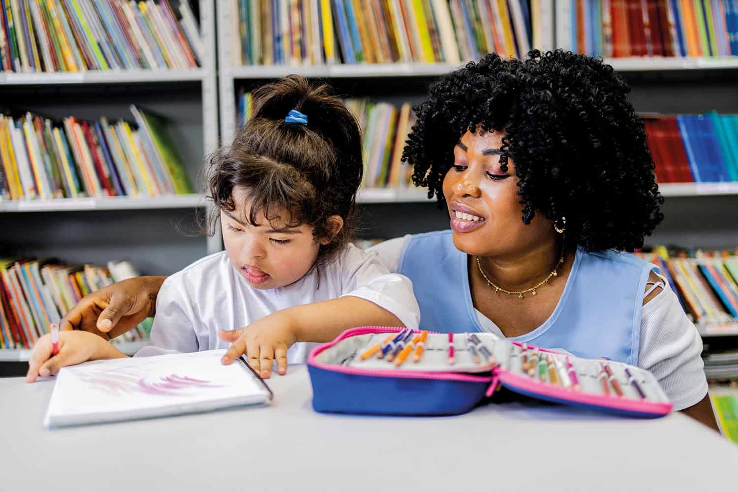 female teacher working with child with learning disabilities who is colouring on a notepad, against a backdrop of shelves of books